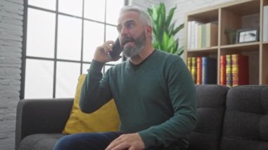 Sıradan yeşil kazaklı, olgun sakallı bir adam geniş pencereli ve kitaplıklı modern dekore edilmiş bir oturma odasından telefon eder..