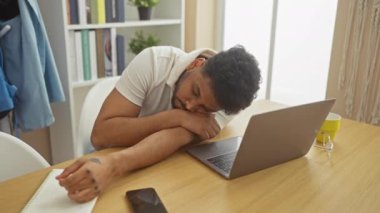 Yorgun bir siyah adam çalışma masasında uykusundan uyanıyor. Evinde parlak bir ofis düzeneği ve arka planda bir laptop ve kahve fincanı var..