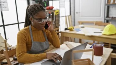 Örgü örgülü Afrikalı Amerikalı kadın marangozluk atölyesinde dizüstü bilgisayar ve telefon kullanıyor..