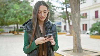 Şehir parkında akıllı telefonuyla mesajlaşan odaklanmış bir kadın, şehir yaşamını ve teknoloji kullanımını gösteriyor..