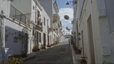 Alberobello, Puglia, İtalya 'da güneşli bir gökyüzünün altında beyazlatılmış evleri gösteren dekor ve çiçeklerle süslenmiş büyüleyici bir sokak..