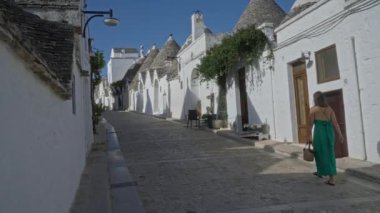 Yeşil elbiseli genç bir İspanyol kadın Alberobello, İtalya 'da beyaz trulli evlerden oluşan büyüleyici bir cadde boyunca yürüyor ve Puglia' daki pitoresk eski kasabayı sergiliyor..