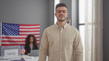 Amerikan bayrağı ve oy verme kabininin olduğu bir yerde kameraya bakan kendine güvenen bir adam, arka planda bir kadın.