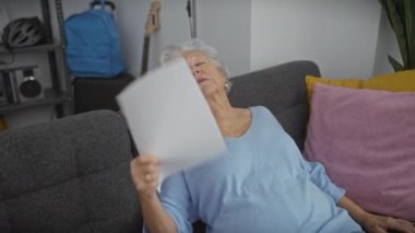 Mavi bluzlu yaşlı kadın gri kanepeye oturup evinde kâğıt sergiliyor, dinlenme ve rahatlama sergiliyor..