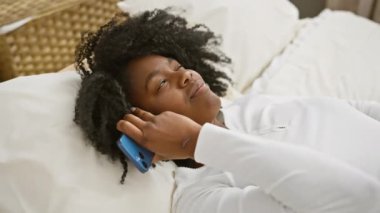 Afro-Amerikalı bir kadın rahat bir yatak odası ortamında akıllı telefonuyla rahatlıyor, evde boş zaman ve teknoloji sergiliyor..