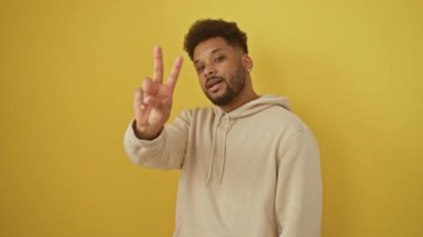 Sarı arka plandaki neşeli Afro-Amerikalı adam zafer işaretini iki parmakla gösteriyor, kendine güvenen bir başarı ve şık kapüşonlu kapüşonlu arkadaşça bir gülümseme yayıyor.