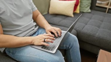 Genç Asyalı adam rahat bir kanepede dizüstü bilgisayar kullanıyor, ev senaryosunda teknoloji tasvir ediyor.