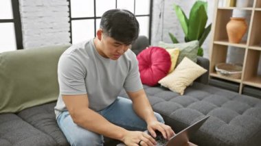 Genç Asyalı bir adam modern bir apartman dairesinde dizüstü bilgisayar kullanıyor, yorgun ve stresli hissediyor..