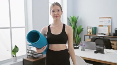 Modern ofis ortamında yoga minderiyle gülümseyen kadın spor ve iş hayatı dengesini gösteriyor.