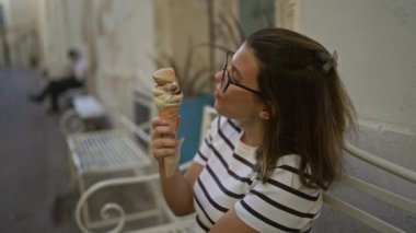 Genç bir kadın güneşli bir günde Lecce, Puglia, İtalya sokaklarında bir bankta otururken büyük bir dondurma külahının keyfini çıkarıyor..