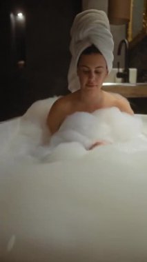 Genç bir kadın köpük banyosunda kafasında havluyla lüks bir banyo ortamında rahatlıyor..