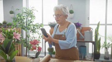 Çiçekçide kıdemli bir kadın çiçeklerin ve bahçe aletlerinin arasında akıllı telefon kullanıyor. Küçük işletmelerde teknoloji sergiliyor..