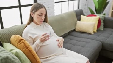 Sakin bir oturma odasında hamile bir kadın renkli yastıklarla kanepede dinlenirken su içer..