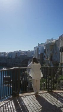 Genç bir kadın polignano manzarasından hoşlanıyor. Puglia, İtalya 'da bir kısrak, manzaralı korkuluklara yaslanıyor. Resimli binalar ve arka planda deniz var..