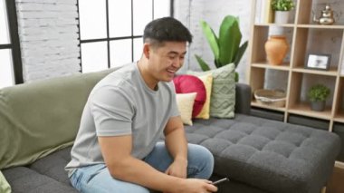 Gülümseyen Asyalı adam, iyi aydınlatılmış bir oturma odasında, elinde uzaktan kumandayla koltukta oturuyor.