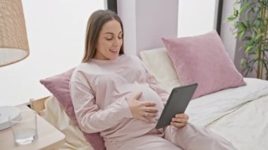 Pembeli mutlu hamile kadın, iyi ışıklandırılmış bir yatak odasında yatakta dinlenirken tablet kullanıyor..