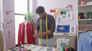 Boynunda bir mezura olan odaklanmış bir terzi dikiş malzemeleri ve mankenlerle dolu parlak bir atölyede tasarımları gözden geçirir..