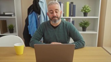 Evde dizüstü bilgisayarla çalışan sakallı, olgun bir adam, esniyor ve modern bir iç mekanda mola veriyor..