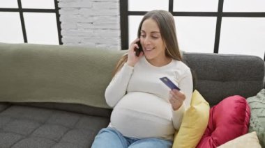 Gülümseyen bir İspanyol hamile kadın telefonda konuşuyor ve elinde bir kredi kartı var..