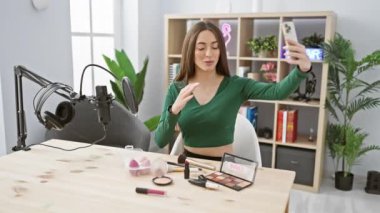 Gülümseyen kadın, makyajlı ve masasında mikrofon olan bir stüdyoda selfie çekiyor..