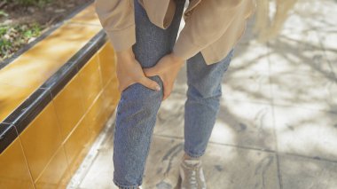 Genç bir kadın dizini açık havada tutuyor, günlük kot pantolon ve bej bir ceket giyiyor, güneşli bir şehir ortamında olası bir rahatsızlık ya da acıyı gösteriyor..