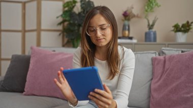 Genç, güzel, sarışın bir kadın mavi bir tablet kullanıyor. İçi bitki ve minderlerle dolu gri bir salonda, rahat bir ev dekorasyonu sergiliyor..