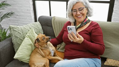 Gülümseyen gri saçlı bir kadın akıllı telefonunu evde rahat bir kanepede nazik köpeğinin yanında kullanmaktan hoşlanır..