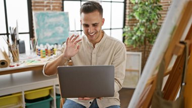 Yakışıklı İspanyol adam, yaratıcı stüdyo ortamında dizüstü bilgisayar başında el sallıyor..