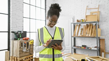 Marangozluk atölyesinde tablet kullanan gülümseyen Afrikalı Amerikalı kadın, marangozluğu ve teknoloji entegrasyonunu resmediyor..