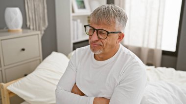 Olgun, gri saçlı, kollarını çapraz yapmış bir adam yatak odasının yatağında oturmuş dalgın dalgın dalgın dalgın dalgın uyuyor..