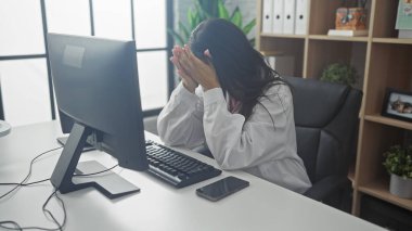 Stressed female doctor in hospital office covers face with hands, showcasing burnout and fatigue at workplace. clipart