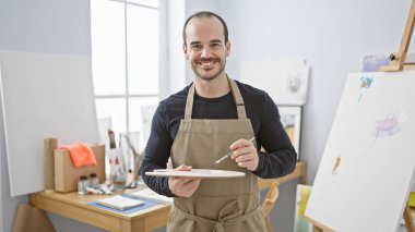 Önlük takan sakallı, gülümseyen kel bir adam bir sanat stüdyosunda palet tutuyor..