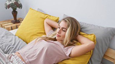 Sarışın genç kadın yatakta rahat bir şekilde uzanıyor, evinde, ellerini başının üstüne koyup dinleniyor, rahat yatak odasının keyfini çıkarıyor, rahatlamak için mükemmel, tam bir sabah uyanıyor..