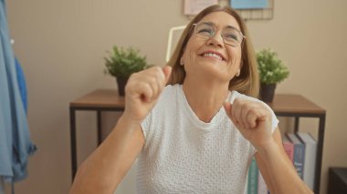 Neşeli olgun bir kadın gözlükleriyle evde dans ediyor, neşe ve canlılığı yansıtıyor.