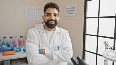 Laboratuvar önlüğü giymiş, sakallı, gülümseyen İspanyol adam, iyi aydınlatılmış bir laboratuarda kollarını kavuşturuyor..