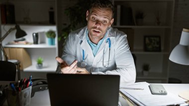 Beyaz önlüklü İspanyol bir erkek doktor, gece loş bir klinikteki video görüşmesinde el kol hareketi yapıyor.