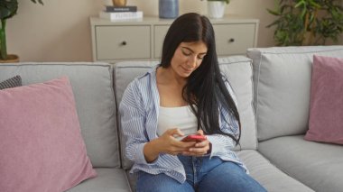Genç bir İspanyol esmer kadın oturma odasında oturuyor, akıllı telefonunu pembe yastıklı rahat gri bir kanepede kullanıyor, rahat bir iç mekan ortamı yaratıyor..