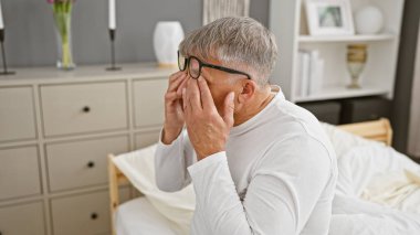 Zor durumdaki gri saçlı bir adam modern bir yatak odasında yatağında oturuyor, yüzünü elleriyle örtüyor..