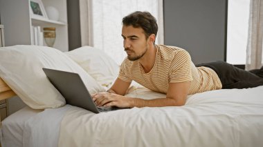Modern yatak odası ortamında dizüstü bilgisayar kullanan sakallı, genç İspanyol bir adam, sıradan bir iş ortamı örneği..