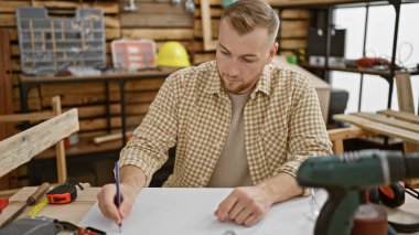 Kareli gömlekli genç sakallı bir adam iyi donanımlı bir marangozluk atölyesinde taslak çiziyor..