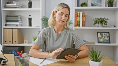 Modern bir ofiste tabletle çalışan beyaz bir kadın. Etrafı bitkiler ve kitaplıklarla çevrili..