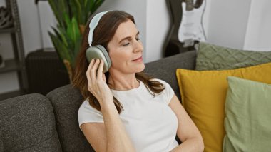 Rahat orta yaşlı bir kadın, rahat bir oturma odasında kulaklıklarla müzik dinliyor..
