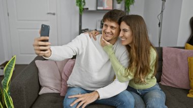 Gülümseyen bir çift sıcak, sevgi dolu bir ev atmosferini yansıtan rahat bir oturma odasındaki kanepede selfie çeker..