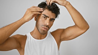 Beyaz kolsuz tişörtlü yakışıklı İspanyol adam izole edilmiş beyaz arka planda saçlarını tarıyor.