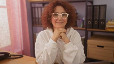 Güzel, orta yaşlı, kıvırcık saçlı ve gözlüklü beyaz bir kadın ofisinde dostça bir gülümsemeyle oturuyor..