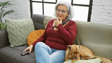 Gülümseyen gri saçlı, gözlüklü bir kadın rahat bir oturma odasında koltukta oturuyor, bir köpeği okşuyor ve uzaktan kumandayı tutuyor..