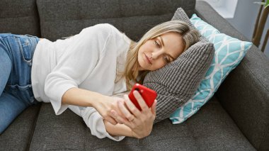 Rahatlamış genç bir kadın evdeki kanepede uzanır, akıllı telefonunu karıştırır..