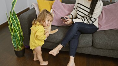 Sarı elbiseli çıplak ayaklı genç bir kız, içi doldurulmuş bir hayvanı kucağında tutuyor. Annesine bakıyor. Annesine bakıyor, gri bir kanepede oturmuş, rahat bir oturma odasında renkli yastıklarla oturuyor..