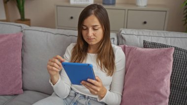 Genç, beyaz bir kadın, oturma odasında tablet kullanıyor, rahat evin içine sorunsuzca karışıyor..