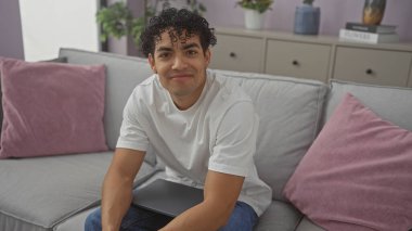 A smiling young hispanic man sitting comfortably on a sofa in a modern living room, exuding casual elegance and warmth. clipart